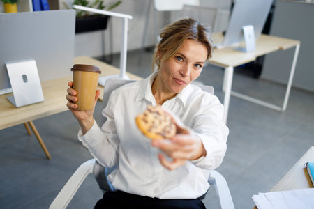 Female office worker with a cup of hot coffee and tasty cookies sits at her workplace in the office