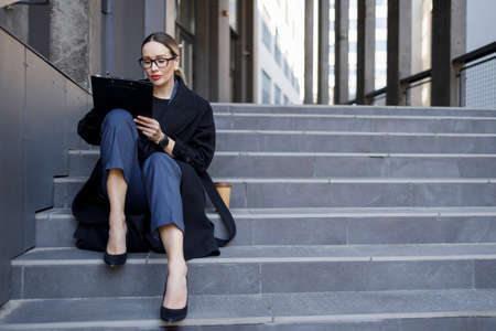 Businesswoman writing on clipboard while sitting on stairs outdoorsの写真素材