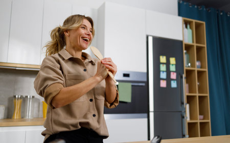 Pretty woman with joyful mood sings in the kitchen while cooking using a wooden spatula as a...