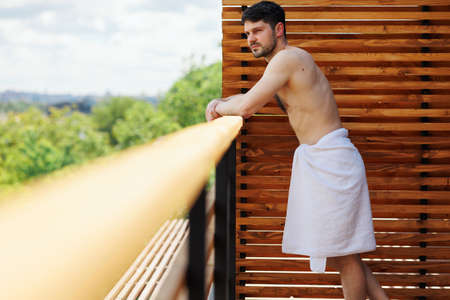 Man wrapped in a towel stands on the balcony of a spa resort leaning against the railingの写真素材