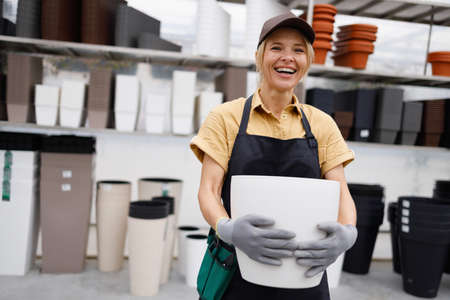 Happy woman in apron and cap holding pot for plant in garden shopの写真素材