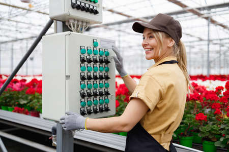 Happy adult woman in apron and gloves pushing buttons on box for controlling watering flowers in the greenhouseの写真素材