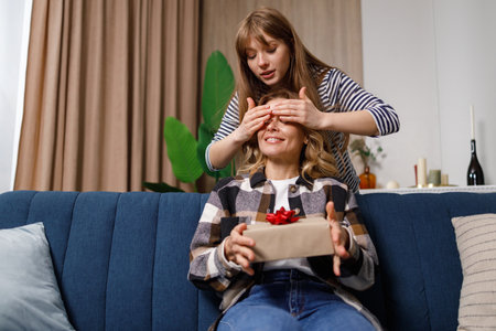 Adult daughter makes an unexpected surprise to her middle aged mom, gives her a gift box to a mothers day or birthday, covering her eyes with hand, sitting on the sofa at homeの写真素材
