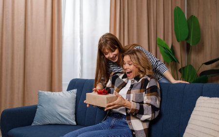 Mother sitting on the couch in the living room is delighted with her daughter's gift. Adult girl congratulates her mom on the holidayの写真素材