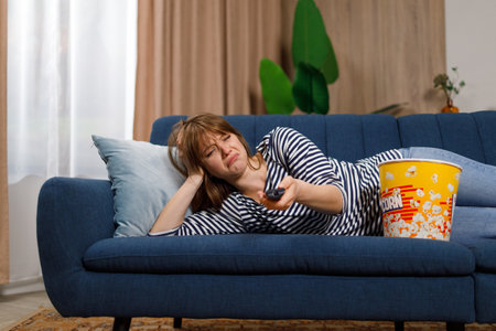 Depressed young woman lies on the couch and switches channels on the TVの写真素材