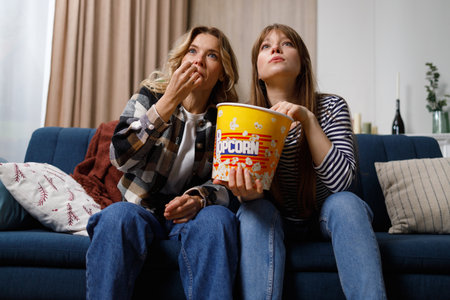 Two women of different ages eating popcorn are concentrated on watching an interesting movie or TV showの写真素材