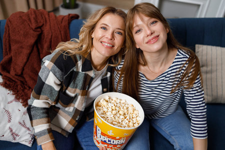 Portrait of mature mother and adult daughter sitting on sofa with bucket of popcorn while watching movieの写真素材