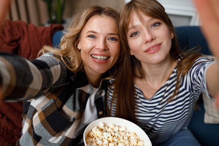 Two woman of different ages take a selfie sitting on the couch with a bucket of popcorn watching a movieの写真素材