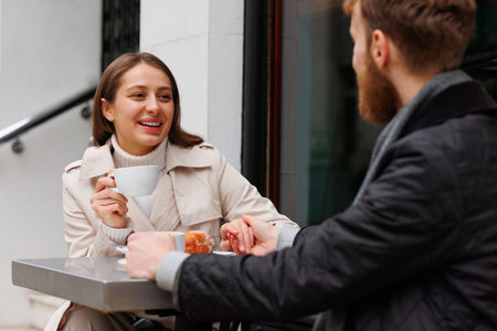 Happy couple drinking coffee and laughing talking to each other sitting at the table of outdoor cafe on city streetsの写真素材