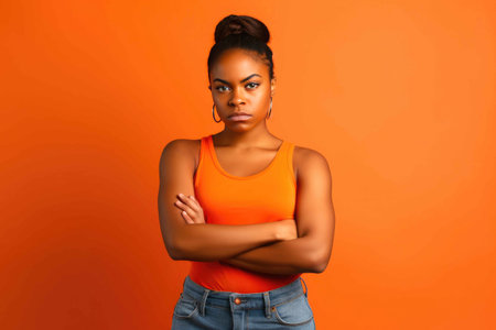 Young black woman wearing a tank top and jeans posing with arms crossed in front of an orange studio background. Generative AIの素材