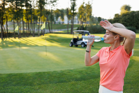 Blonde Golfer Hydrating on Course, Golf Cart in Scenic Viewの写真素材