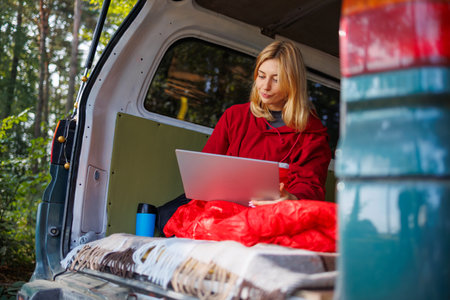 A young woman works on a laptop while sitting in her van in the forest. Work away from homeの写真素材