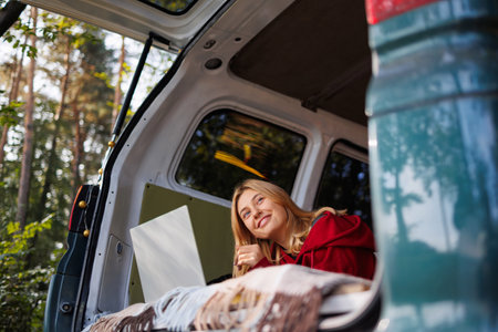 Joyful young woman lies in a camper van in the forest with a laptop and looks away through the tailgateの写真素材
