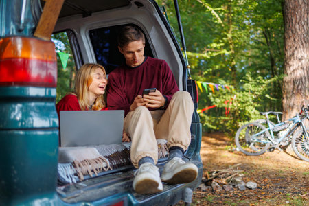A young couple while relaxing in the forest in a homemade camper van has fun watching something on a...