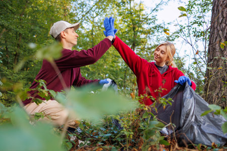 Motivated by care for the environment, young man and woman high-five each other while cleaning the forest from garbage waste. Green activists cleaning up natureの写真素材