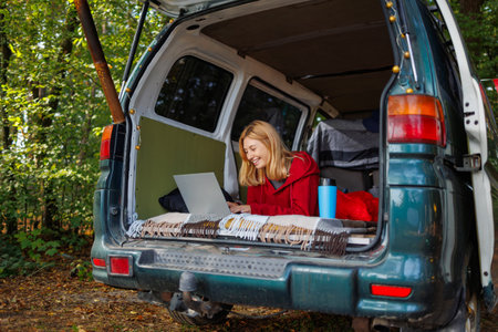 Happy young woman lying in homemade camper van with laptop in forestの写真素材