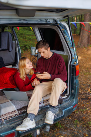 Young couple watching something on a smartphone while sitting in a homemade camping van while relaxing in the forestの写真素材