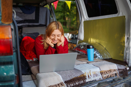 Joyful young woman lying in van looking at laptop while relaxing in campingの写真素材