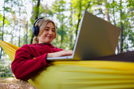 A girl wearing headphones lies on a hammock in the forest and uses a laptopの写真素材