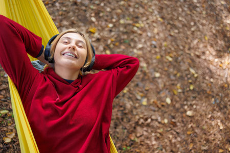 Cheerful girl in a red hoodie with headphones is chilling on a yellow hammock in the forestの写真素材