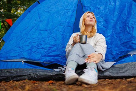 A young positive woman with a camping cup in her hands sits in a tent and looks at the forest aroundの写真素材