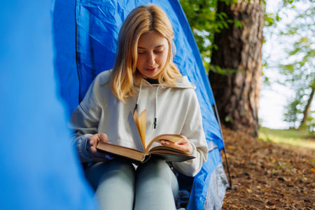 Young woman leafing through a book while sitting in a tent surrounded by natureの写真素材