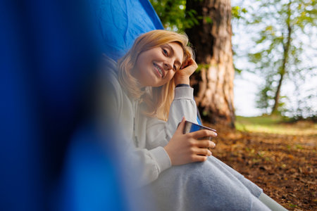 Young woman sitting at the entrance to a tent with a mug in her hand and looking at the cameraの写真素材