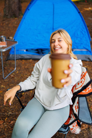 A girl sitting in a camping chair against the backdrop of a tent in the forest holds out a mug of coffee to the cameraの写真素材