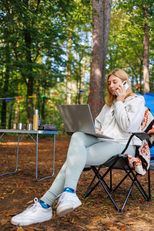 Young woman talking on the phone while sitting in a chair with a laptop in a forest campingの写真素材