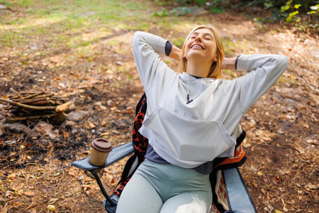 A young woman in a good mood is sitting on a chair relaxing during a camping tripの写真素材
