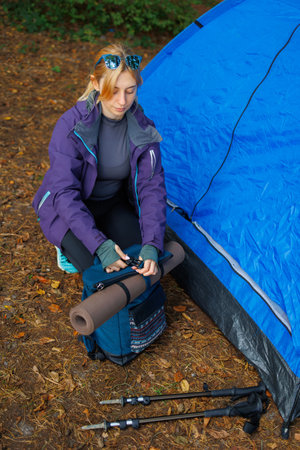 Woman in the forest near a tent attaches a yoga mat to a backpack before going on a hikeの写真素材
