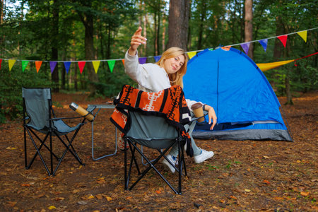 Young woman sitting on a chair at a forest campsite calling to join herの写真素材