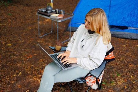 Happy young girl with a laptop is sitting in a camping chair in the middle of the forest with a tent in the backgroundの写真素材