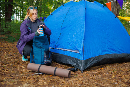 Female tourist near a tent in the forest puts a thermal mug in her backpack getting ready for a hikeの写真素材