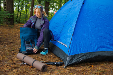 Young woman tourist packing backpack near tent in forest campingの写真素材