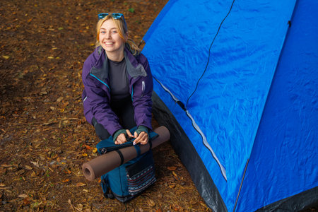 Happy female hiker near the tent in the forest preparing for a hike by packing and attaching a yoga mat to a backpackの写真素材