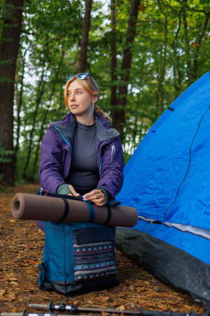 Girl in the forest near a tent is preparing for a hike. Female hiker attaching a yoga mat to a backpackの写真素材