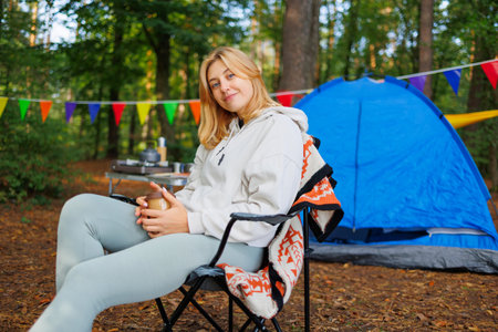 Portrait of a young woman sitting on a chair with a cup of coffee in a forest camping with a tent and carnival garlands on the backgroundの写真素材