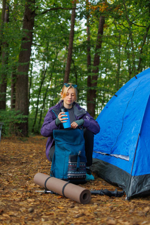 A woman puts a thermal mug in her backpack while going on a hike after spending the night in a tent in the forestの写真素材