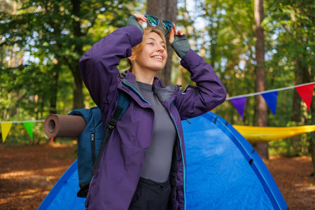 Happy female tourist with a backpack in the forest near the tent puts on sunglassesの写真素材