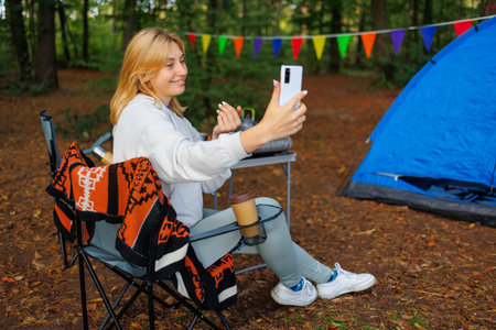 A young woman vacationing at a forest campsite talks on video via a smartphone and asks to join herの写真素材