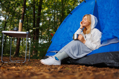 Woman with closed eyes and cup in hands enjoying nature while sitting in tent and drinking hot drinkの写真素材
