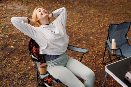 Young female tourist stretching while sitting on a chair near a tent in a forest camping. Enjoyment of rest in natureの写真素材
