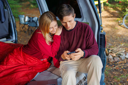 Cute couple watching something interesting on a smartphone while sitting in their homemade camping van in the forestの写真素材