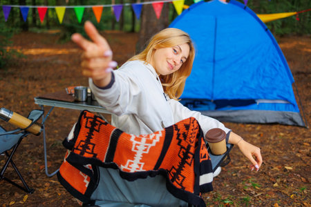 A female tourist sitting on a chair in a forest camping shows a hand gesture to joinの写真素材
