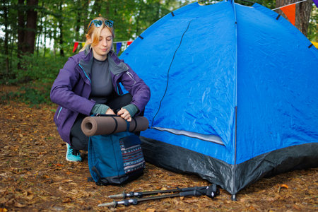 Young woman near a tent in the forest prepares for a hike, attaches a yoga mat to a backpackの写真素材