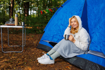 A cute young woman is enjoying the nature of the forest sitting in a tent with a mug of hot drinkの写真素材