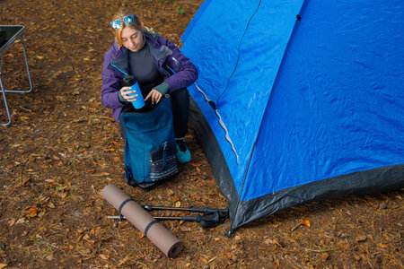 Top view of tourist woman near a tent in the forest puts a thermal mug in her backpack getting ready for a hikeの写真素材