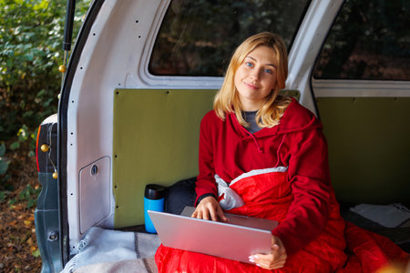 A beautiful red-haired girl sits in a van with a laptop during a vacation in natureの写真素材