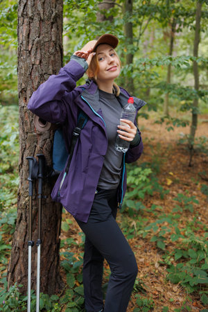 Female hiker adjusting cap while holding water bottle and smiling in forestの写真素材